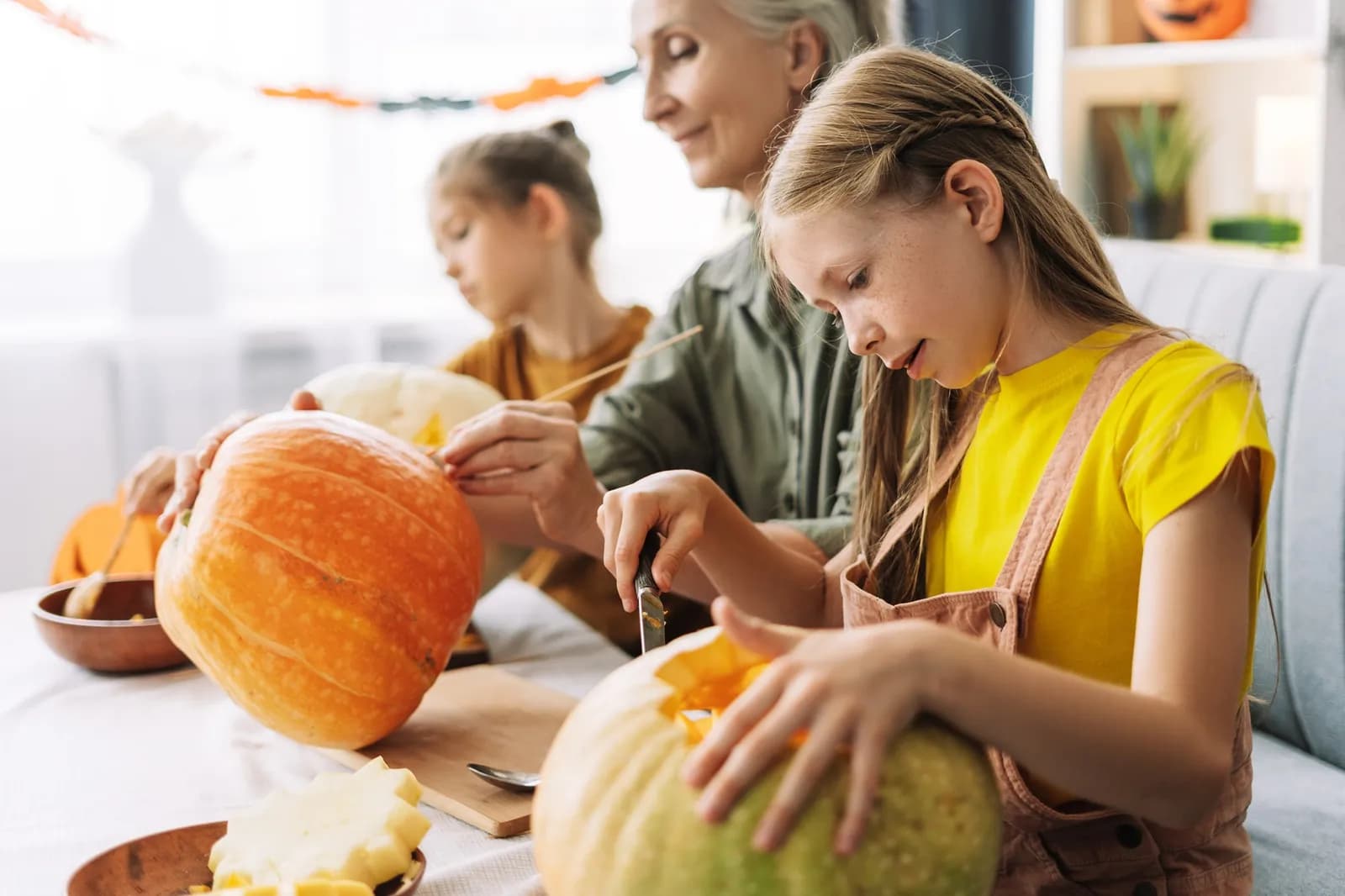 A young girl in a yellow shirt carving a pumpkin with an older woman and another child in the background, all preparing for Halloween.