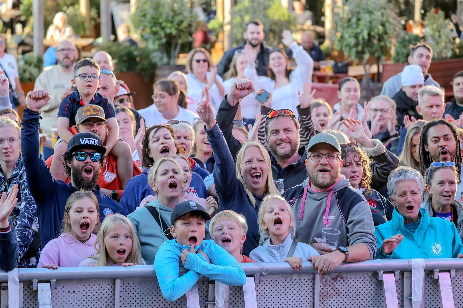 A diverse crowd of people, including many children, are cheering and looking upwards, many with open mouths, as if watching a performance or event.