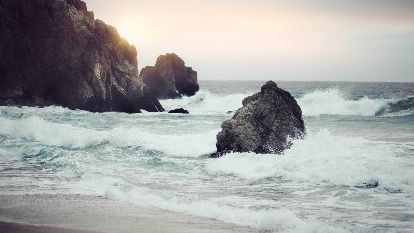 Waves crashing against large gray rocks on a cloudy day.