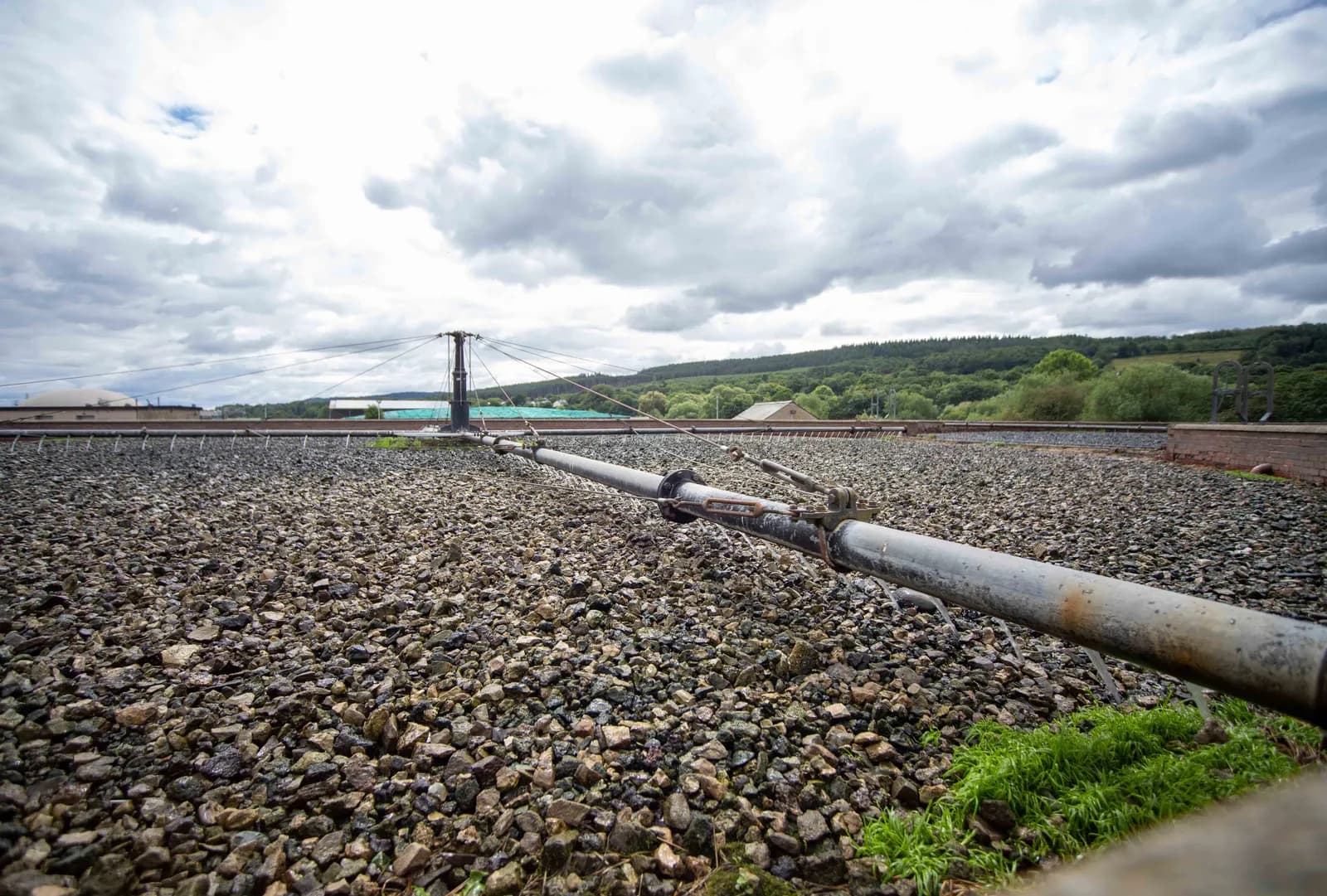 A large pipe extends over a vast bed of gravel under a cloudy sky, with green hills and structures visible in the distance.