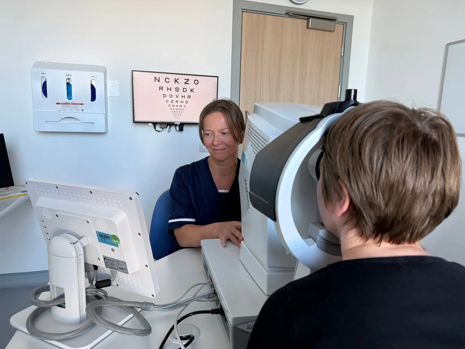 A medical professional in blue scrubs looks at a patient using an eye examination machine, with a vision chart visible in the background.