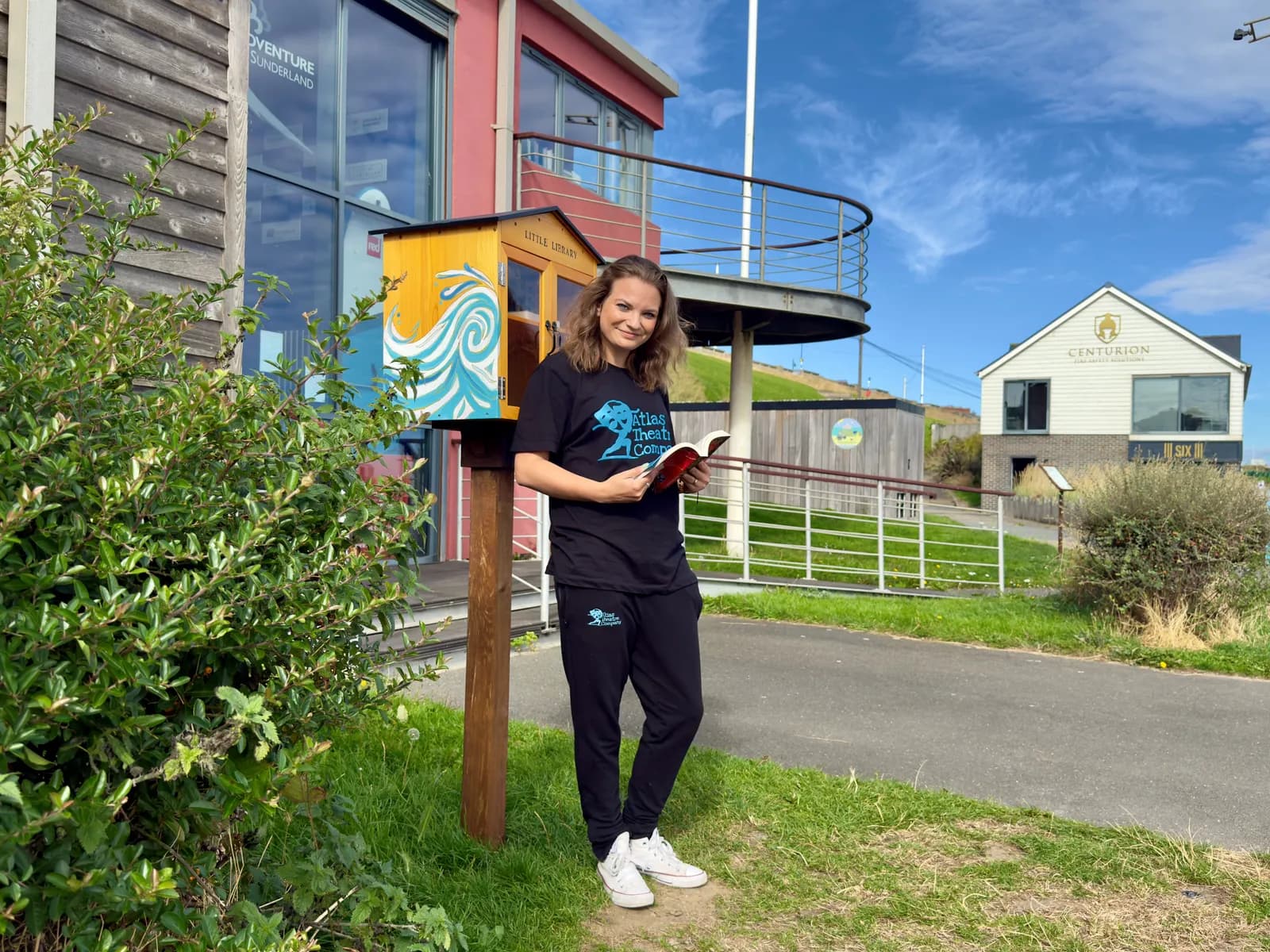 A woman in a black t-shirt and black pants stands next to a small, yellow, wave-painted bookshelf, holding an open book, with buildings and a blue sky in the background.