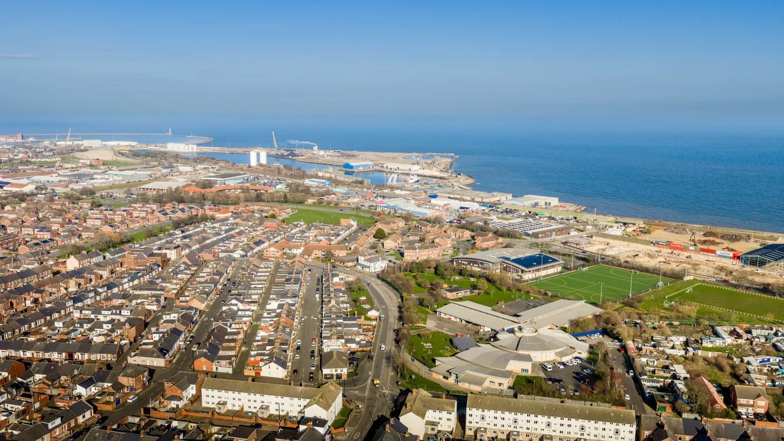 An aerial view of a coastal town showing rows of residential houses, a sports field, industrial buildings, and a harbor extending into the blue sea under a clear sky.